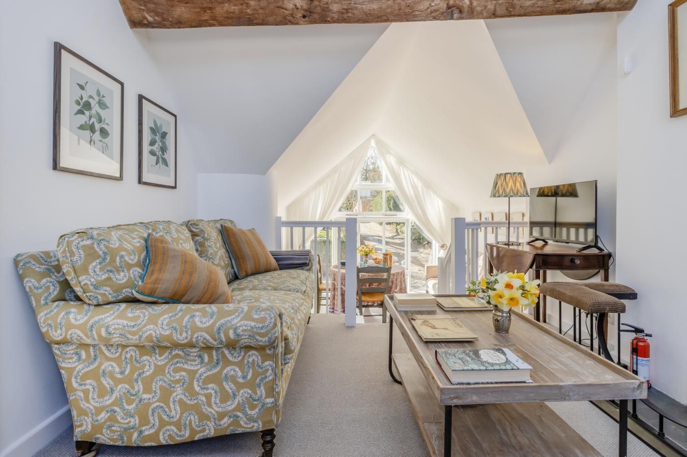Ein gemütliches Zimmer mit einer gemusterten Couch, einem Holztisch mit Buch und Blumen sowie einem großen Fenster mit Blick ins Freie.