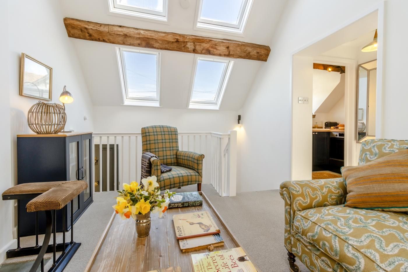 A cozy loft apartment with a high ceiling, skylights, a plaid armchair, a sofa, a small table with books, and a radiator.