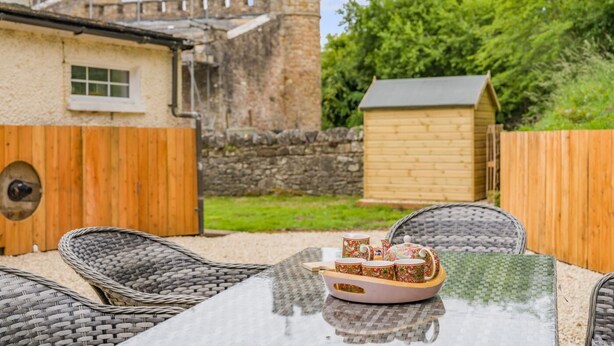Eine Terrasse mit Korbstühlen und einem Tisch, einem Holzzaun und einer Steinmauer mit einem Turm im Hintergrund.