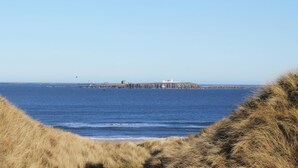 A coastal landscape with a lighthouse on a small island, surrounded by calm sea and grassy dunes. - 2 Bed in Seahouses (oc-gd1314) (Seahouses)