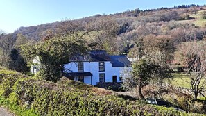 A white building with a dark roof, surrounded by trees and a hilly landscape.