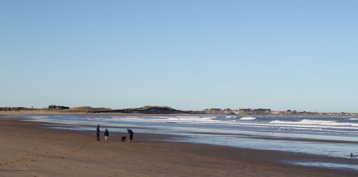 Ein weiter Sandstrand mit sanften Wellen, einigen Personen und Hügeln im Hintergrund.