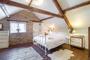 A bedroom with a stone wall, wooden beams, a white bed, a nightstand, a dresser, and a window with a view. - 3 Bed in Llanvetherine (oc-g31918) (Llanvetherine)