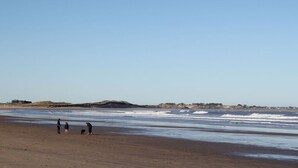 Une vaste plage de sable fin, avec de douces vagues, quelques personnes et des collines qui se dessinent à l’horizon.