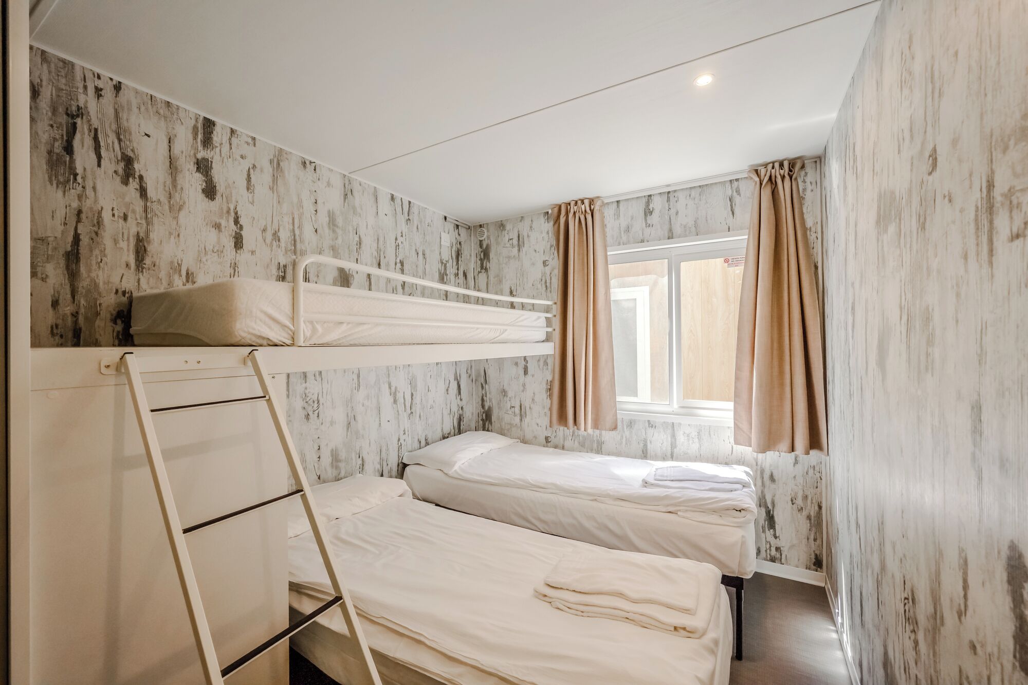 A bunk bed room with a ladder, a window with curtains, and a wooden wall.