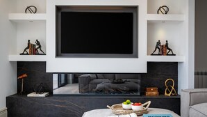 Uma sala de estar moderna com uma televisão de tela grande montada acima de uma lareira, um sofá confortável e uma mesa de centro com livros e uma fruteira.