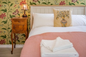 Ein ordentlich bezogenes Bett mit weißen Laken und einer rosa Decke, ein Nachttisch aus Holz mit einer Lampe und eine Wand mit floralem Muster.