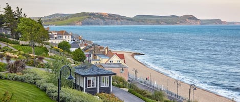 Un pueblo costero con un paseo marítimo empedrado que lleva a una playa, casas con techos rojos y un faro a lo lejos.