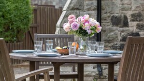 A round wooden table set for four with plates, glasses, and a bowl of fruit. A vase with pink and white flowers is on the table. - 4 Bed in Bamburgh (oc-gd1264) (Bamburgh)