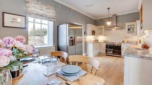 A modern kitchen with white cabinets, stainless steel appliances, and a dining area with a table set for two. - 4 Bed in Bamburgh (oc-gd1264) (Bamburgh)