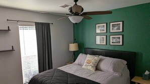 Una camera da letto con un letto, una ventola a soffitto, una finestra con tende e quadri appesi al muro.
