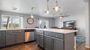 Una cucina moderna con mobili grigi, lavastoviglie, luci a sospensione, una ventola a soffitto e un'ampia finestra che dà su un giardino.