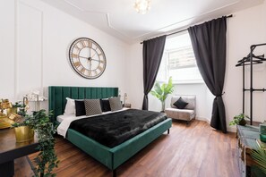 A modern bedroom with a green upholstered bed, a round wall clock, a side table with decorative items, and a window with curtains. - 3 Bedrooms Termini Apartment (Roma)