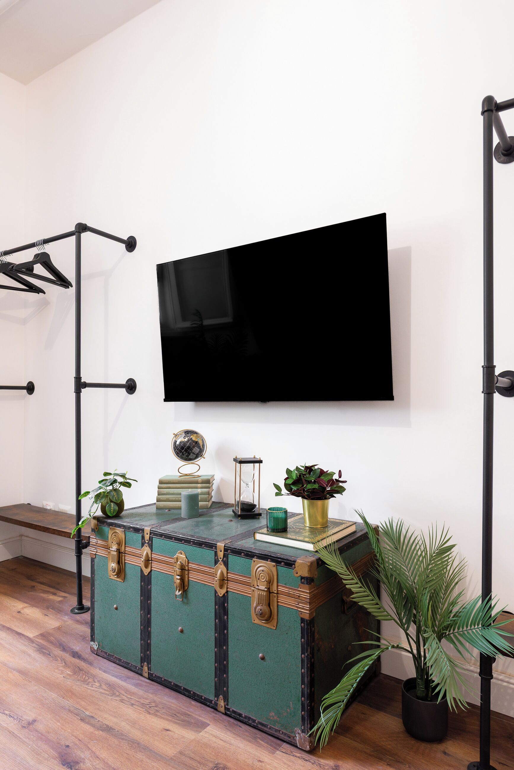 Una stanza con un baule verde, una TV a parete, un appendiabiti e una pianta in vaso.