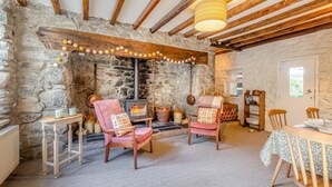 Une pièce chaleureuse avec un mur en pierre, des poutres en bois, une cheminée, deux fauteuils, une table et une bibliothèque.