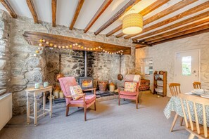 Ein gemütliches Zimmer mit Steinwand, Holzbalken, Kamin, zwei Stühlen, einem Tisch und einem Bücherregal.
