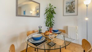 A dining area with a round glass table, four chairs, a potted plant, a framed picture, and a radiator.
