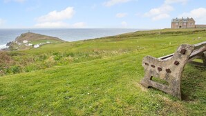 A grassy landscape with a stone bench, a distant building, and a view of the sea.