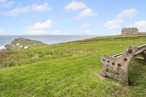 A grassy landscape with a stone bench, a distant building, and a view of the sea.