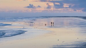 Una playa con un mar tranquilo, dos personas caminando y un perro.