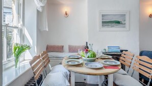 A cozy dining area with a round wooden table set for four, surrounded by wooden chairs with white cushions. A vase with white flowers is on the windowsill, and a framed picture of a beach scene hangs on the wall. - 3 Bed in St. Teath (oc-r31945) (Saint Teath)