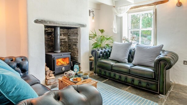 A cozy living room with a stone fireplace, a black leather sofa, and a window with a view of greenery. - 3 Bed in St. Teath (oc-r31945) (Saint Teath)