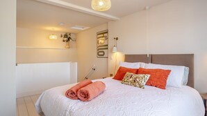Een slaapkamer met een groot bed, witte beddengoed en een hoofdeinde. Er staat een nachtkastje met een lamp en een wandplank met bloemen.