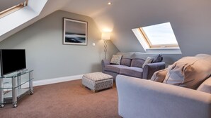 A cozy loft bedroom with a sloped ceiling, a large window, a flat-screen TV, a sofa, and a small ottoman. - 4 Bed in North Sunderland (oc-gd0238) (North Sunderland)