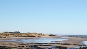 A coastal landscape with a sandy beach, rocky outcrops, and a distant castle-like structure. - 4 Bed in North Sunderland (oc-gd0238) (North Sunderland)