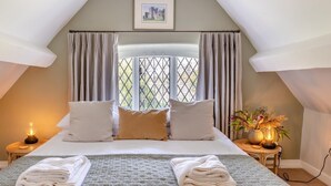 Ein Schlafzimmer mit Bett, zwei Nachttischen, einem Fenster mit Vorhängen, einem gerahmten Bild an der Wand und einem Beistelltisch mit Lampe und Blumenvase.
