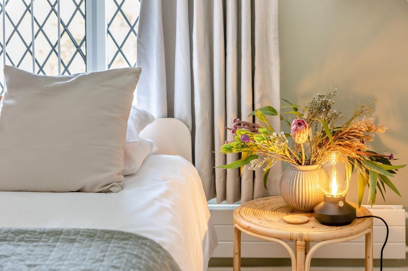 A neatly made bed with pillows, a bedside table with a vase of flowers and a lamp, and a window with curtains.