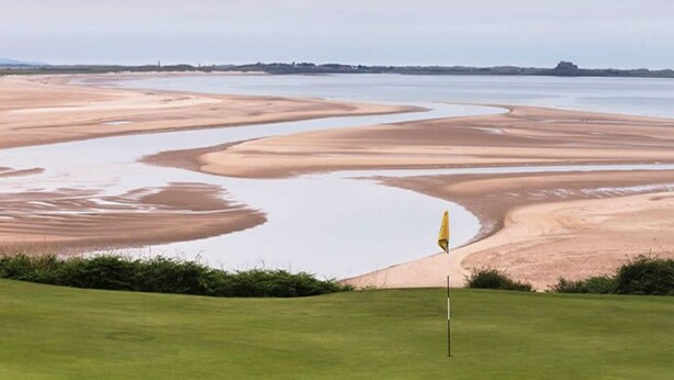 Ein Golfplatz mit einer Fahne im Vordergrund und einer großen Wasserfläche im Hintergrund.