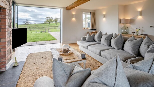 Un salon moderne avec un grand canapé gris, un téléviseur à écran plat et une vue sur un paysage verdoyant, visible à travers une porte vitrée.