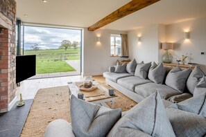 Un salon moderne avec un grand canapé gris, un téléviseur à écran plat et une vue sur un paysage verdoyant, visible à travers une porte vitrée.