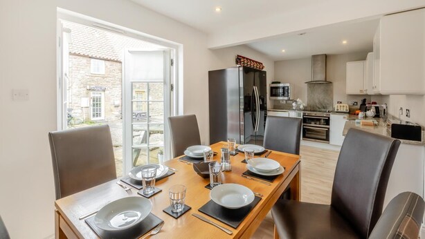 A dining area with a table set for four, a kitchen in the background, and a view of a stone building outside. - 3 Bed in Belford (oc-gd0144) (Adderstone with Lucker)