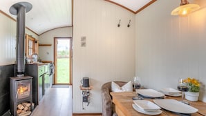 Une cuisine chaleureuse avec un poêle à bois, un coin repas avec une table dressée pour deux personnes, et une vue sur le jardin par une porte ouverte.