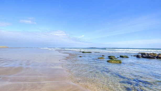 Een strand met helder water, rotsen en een verre kustlijn.