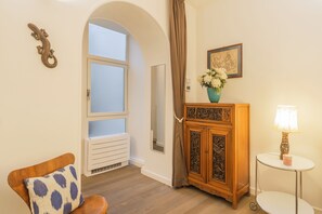 Ein Zimmer mit einem Holzschrank, einem kleinen Tisch mit Lampe, einem Sessel mit gemustertem Kissen und einem Fenster mit Vorhang.
