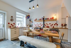 A modern kitchen with white cabinets, a wooden dining table, and a bench. There are hanging lights, a window, and a colorful wall hanging. - 3 Bed in Hawes (oc-ds959) (Hawes)