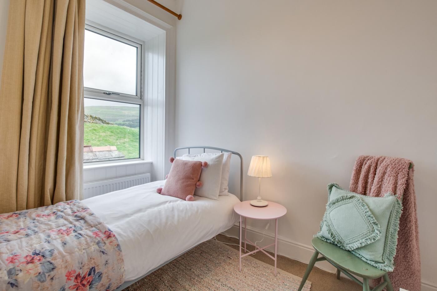 A bedroom with a bed, a window, a bedside table with a lamp, and a chair with a blanket.