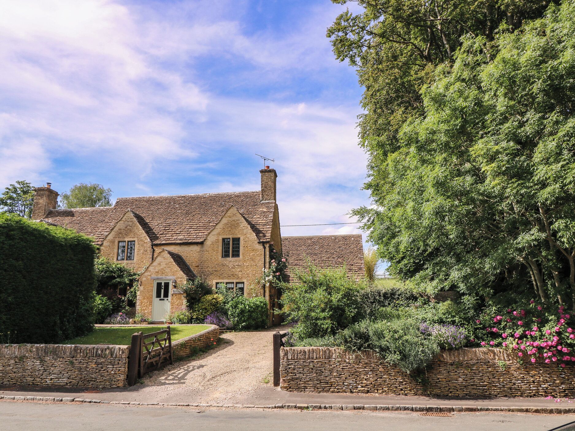 A traditional stone house with a gravel driveway, surrounded by lush greenery and a stone wall.