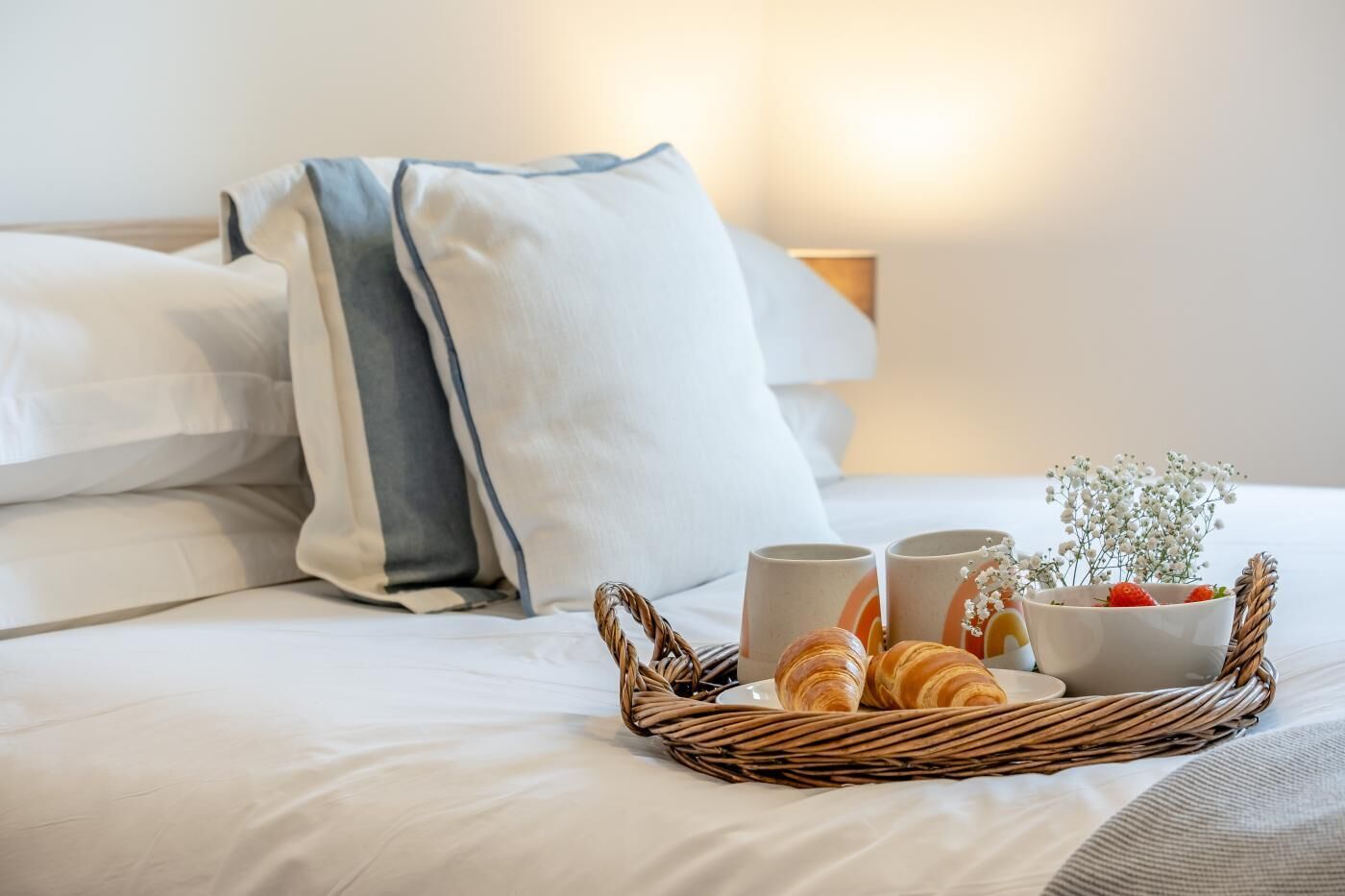 A neatly made bed with white linens and a grey and white pillow. A wicker tray with croissants, two mugs, and a small bowl with flowers is placed on the bed.