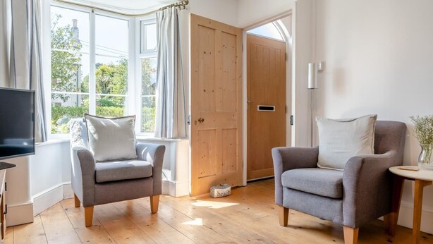 Ein Zimmer mit zwei grauen Sesseln, Holzboden, Fernseher und einem großen Fenster mit Vorhängen.
