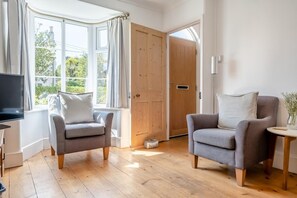 Quarto com duas poltronas cinzentas, piso de madeira, televisão e uma grande janela com cortinas.