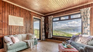 A cozy room with wooden walls and floors, two sofas facing a large window with a scenic view, and a small table with cups and a teapot. - 3 Bed in Barmouth (oc-c31942) (Barmouth)