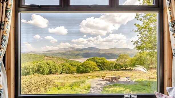 A window with a view of a grassy landscape, a wooden table, and two glasses on the windowsill. - 3 Bed in Barmouth (oc-c31942) (Barmouth)