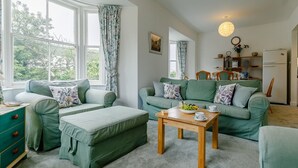Ein gemütliches Wohnzimmer mit grünen Sofas, einem Holztisch und Blick ins Freie durch große Fenster.