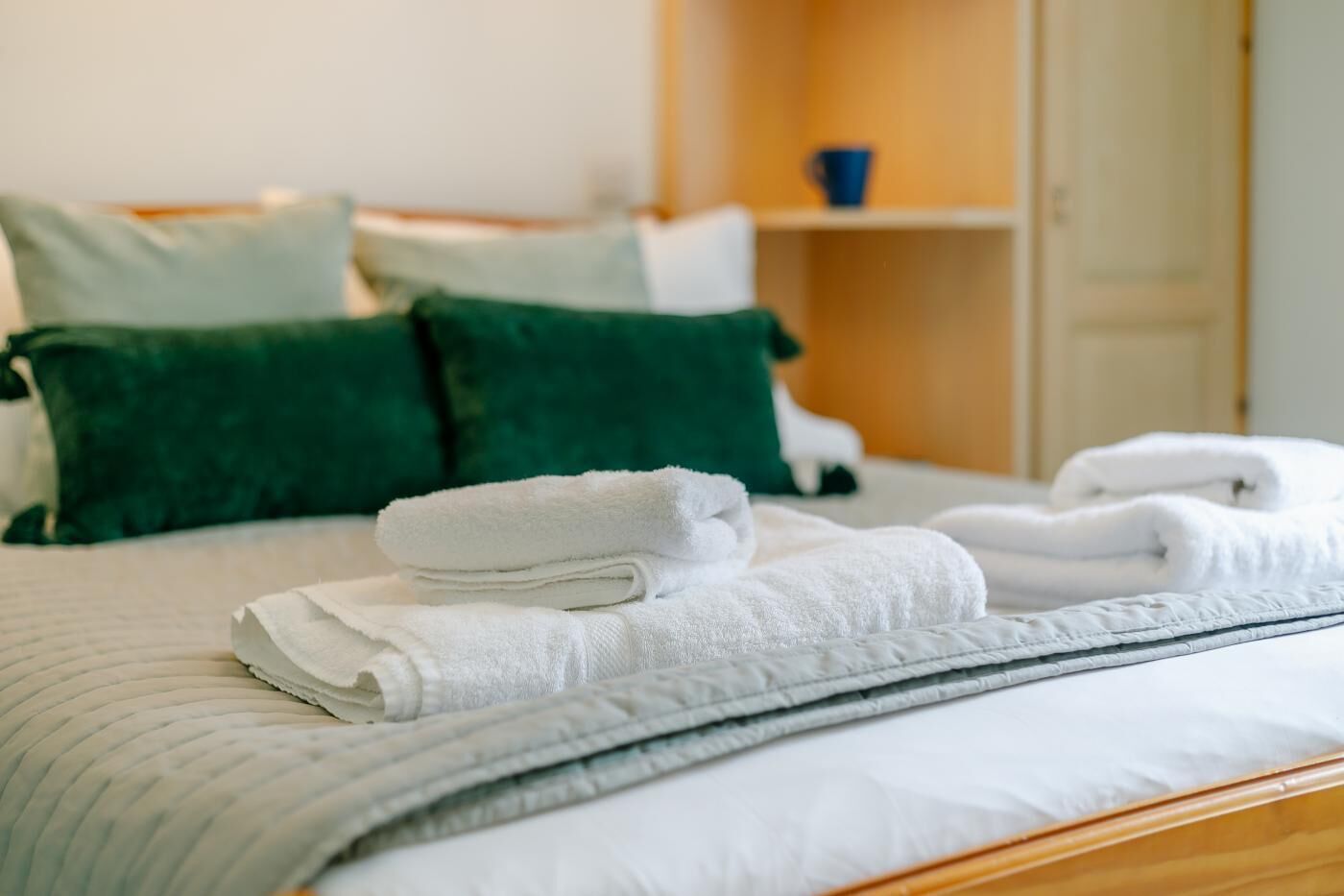 Un lit bien fait, avec des oreillers blancs et verts, une serviette blanche pliée et une serviette enroulée posées sur un couvre-lit blanc.