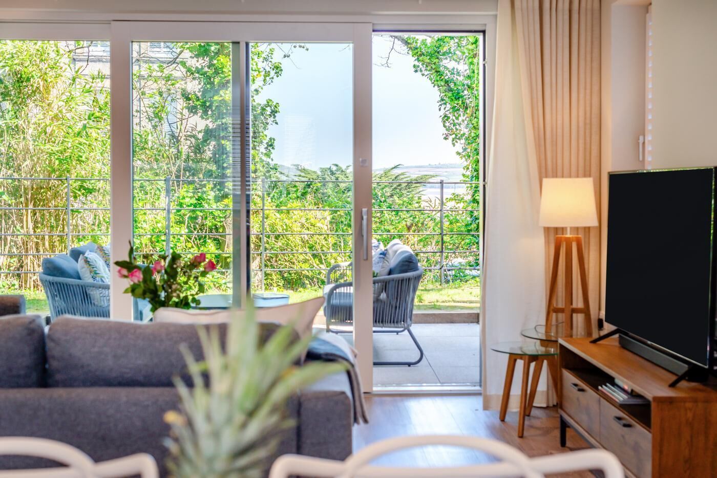 Een moderne woonkamer met een bank, stoelen, een televisie en uitzicht op groen via grote schuifdeuren.