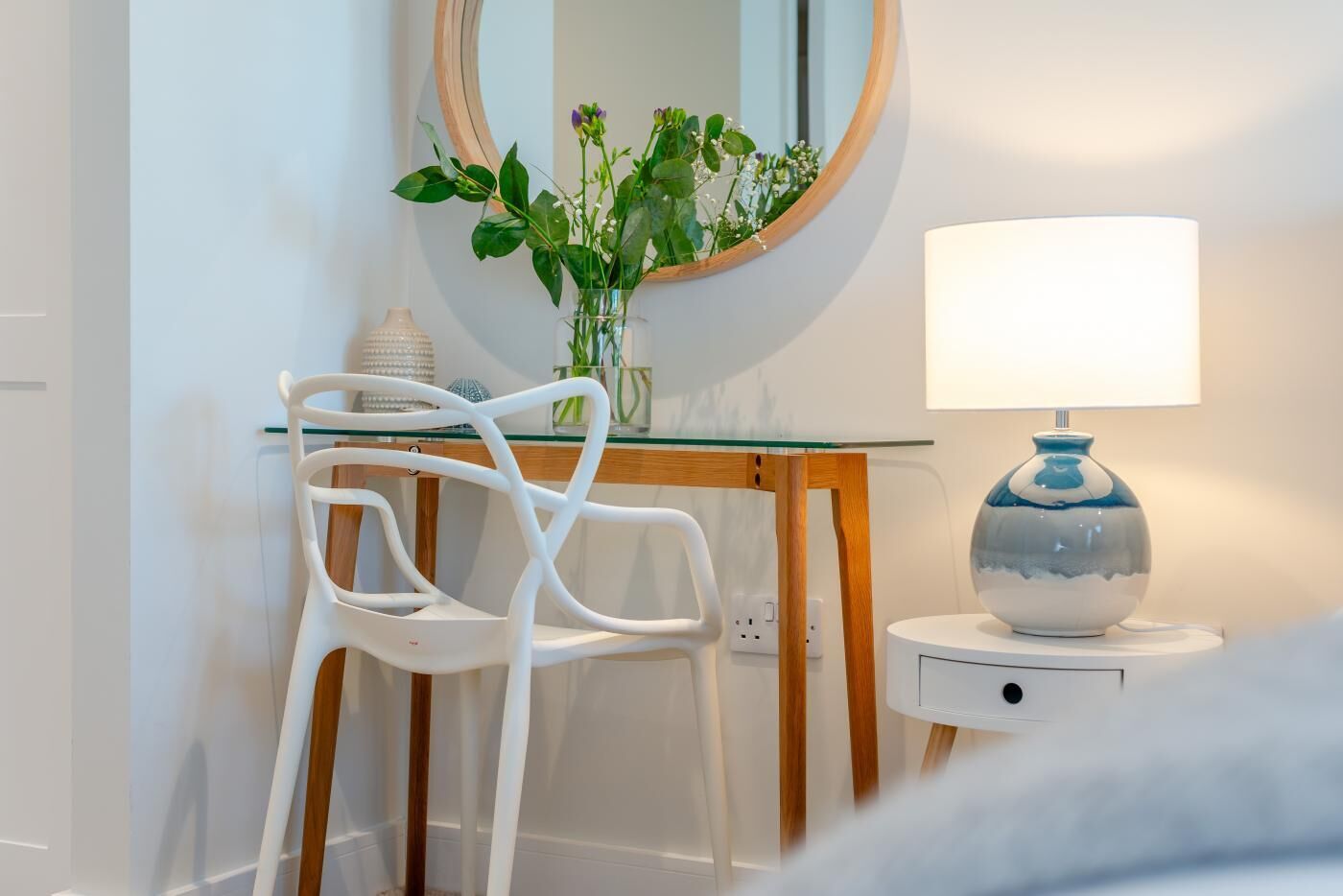 Een moderne kamer met een witte stoel, een glazen tafel, een houten bijzettafeltje met een lamp en een spiegel met houten lijst.